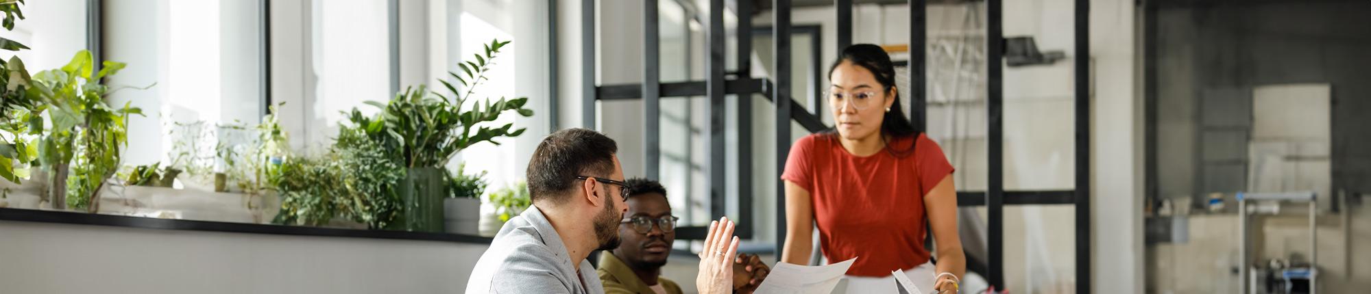 Two colleagues seated at a table engaging in conversation with a colleague standing nearby